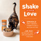 Calico cat eating Instinct Raw Boost Shakers cat food topper from a beige bowl, next to the product bottle.
